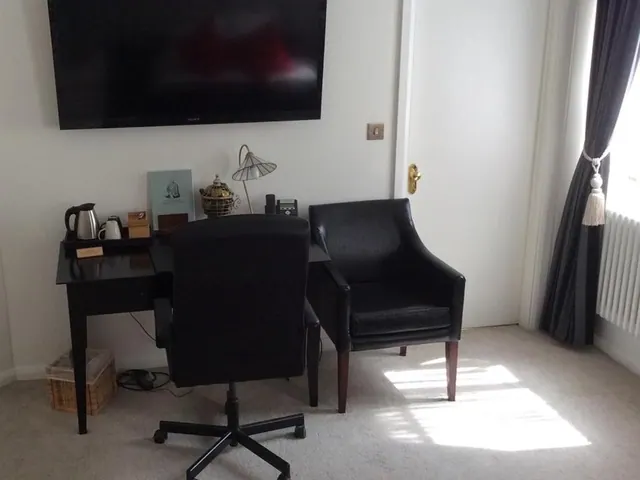 A hotel room corner with a wall-mounted Sony television above a black desk holding a kettle, a lamp and a book, with a black office chair and a black leather armchair in front of a white door and curtained window.