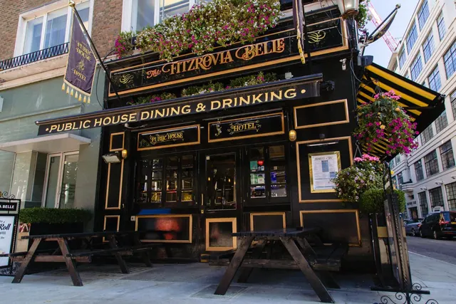 The street facade of The Fitzrovia Belle pub and hotel, with black paintwork, gold lettering, hanging flower baskets and wooden picnic benches on the pavement.