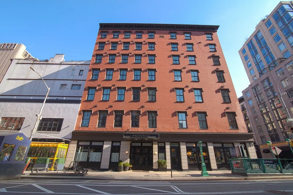The red brick front of the hotel building, with its FREDERICK sign above the entrance and a subway entrance on the street beside it, under a clear blue sky.