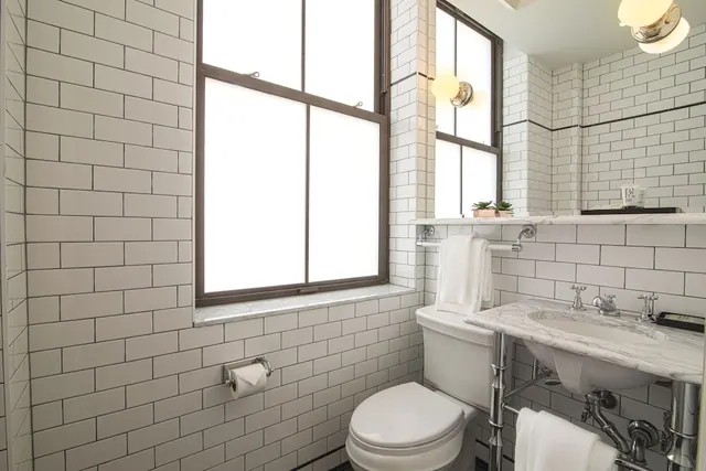 A white subway-tiled bathroom with a marble-topped pedestal sink, a toilet, folded towels and a tall window with frosted glass.