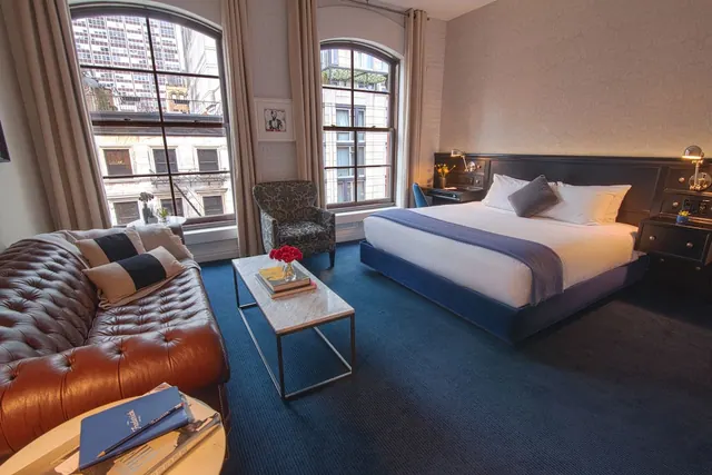A bedroom with a king bed, a tufted brown leather sofa, an armchair and a coffee table, with arched windows overlooking city buildings.