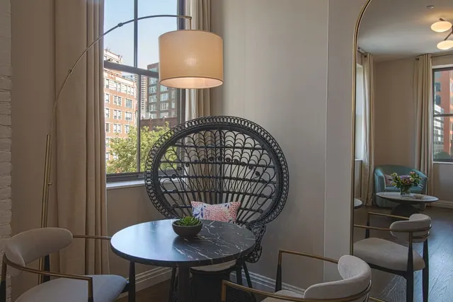 A corner of a hotel room with a round marble table, a black wicker peacock chair and an arc floor lamp beside a window looking onto brick buildings, with a mirror reflecting another seating area.