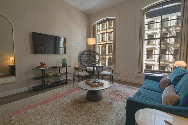 A furnished living room with a teal sofa, a wall-mounted television, a round coffee table and tall arched windows looking onto a neighbouring brick building with fire escapes.