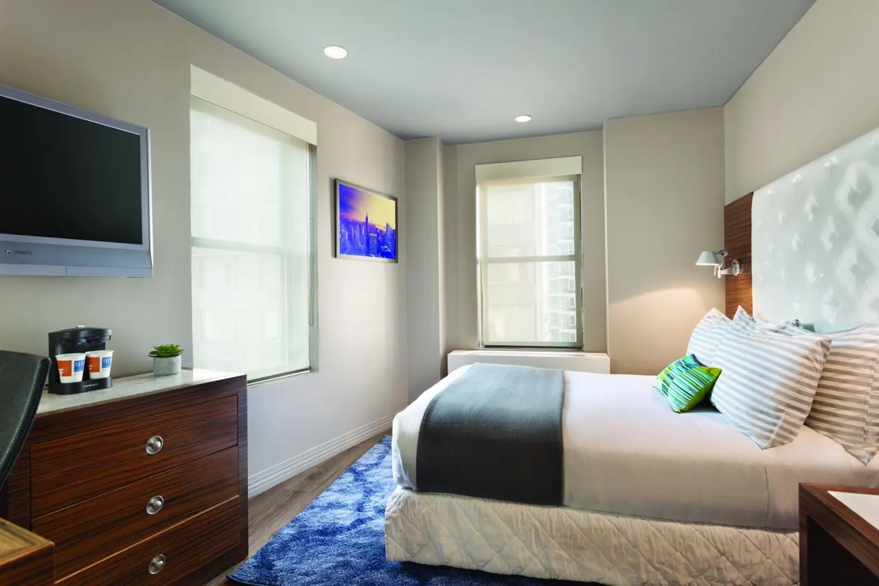 A hotel bedroom with a made double bed, a wall-mounted flat-screen TV, a wooden dresser with a coffee maker, and two windows with shades.