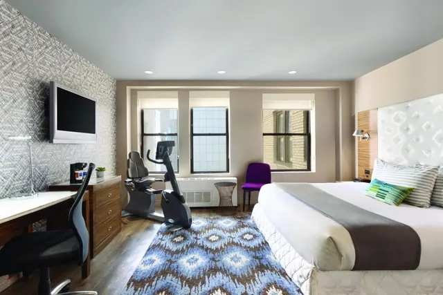 A hotel bedroom with a king bed, a wall-mounted television, a desk, an exercise bike and a purple chair in front of three windows looking onto neighbouring buildings.