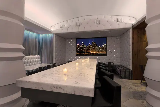 A long marble dining table with lit candles and black leather chairs sits in a room with tufted white walls and a wall-mounted television showing a city skyline at night.