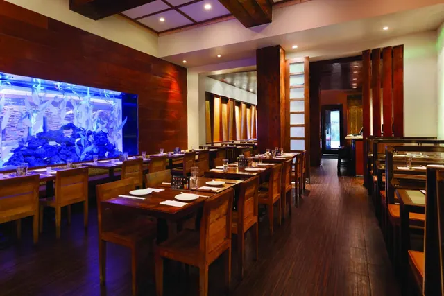 A dimly lit restaurant dining room with wooden tables and chairs, and a large illuminated aquarium built into the wall on the left.