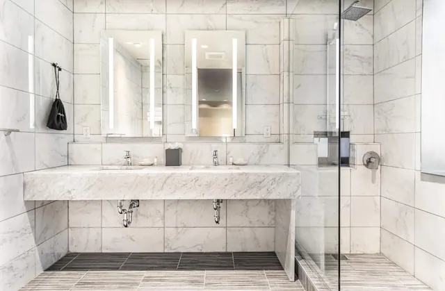 A bathroom with marble-tiled walls, a floating double marble vanity with two illuminated mirrors, and a glass-enclosed walk-in shower.