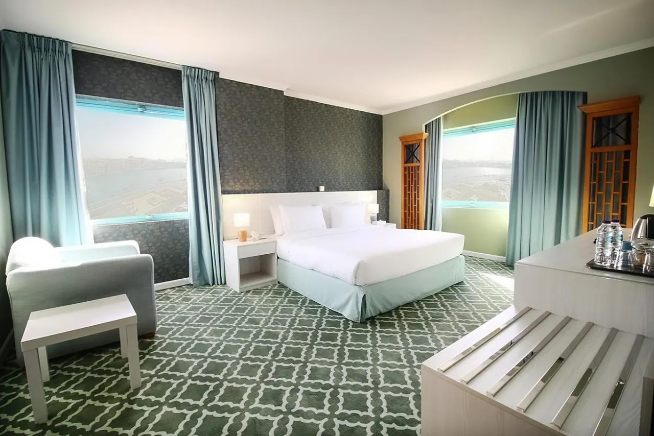 A hotel bedroom with a king bed, an armchair, a bedside table, and windows looking out over water and distant buildings.