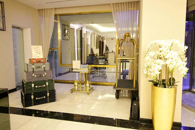 A hotel lobby with stacked luggage trunks, a gold luggage trolley, a gold drinks trolley and a gold planter of white orchids on a marble floor.