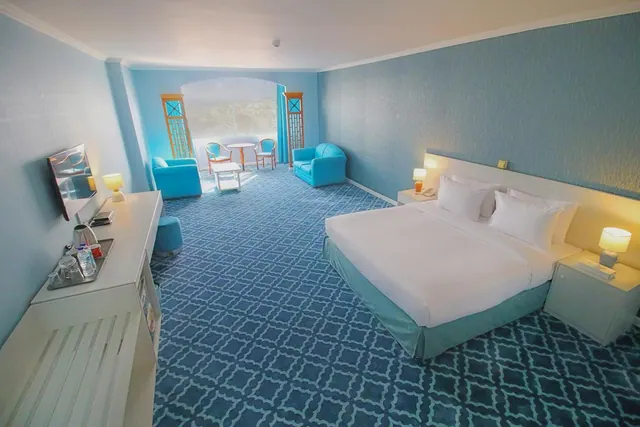 A blue-themed hotel bedroom with a king bed, two armchairs, a wall-mounted television, and a kettle and glasses on a cabinet above a fridge, with a printed city backdrop beyond the arched doorway.