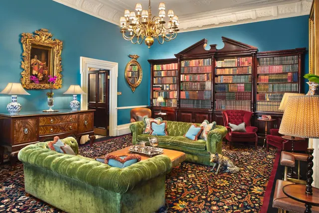 A lounge with teal walls, two green velvet chesterfield sofas, a wall of bookcases, a chandelier and a bronze fox sculpture on a patterned rug.