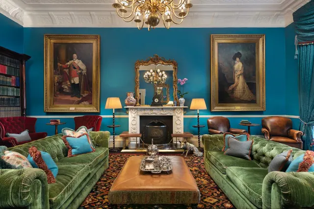 A lounge with teal walls, green velvet sofas, framed portraits, a white marble fireplace and a brass chandelier.