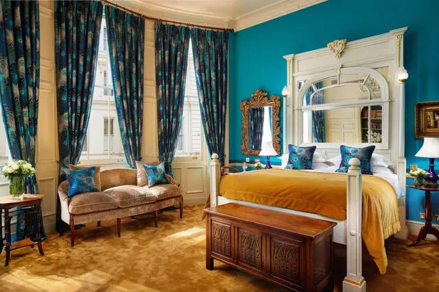 A bedroom with teal walls, peacock-feather curtains, a double bed with a gold throw, a velvet sofa and a carved wooden chest at the foot of the bed.
