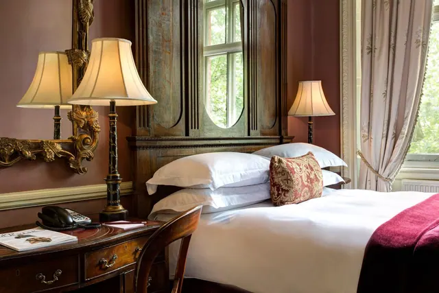A bedroom with a made double bed, a carved wooden headboard, table lamps, a writing desk with a telephone, and a window showing trees outside.