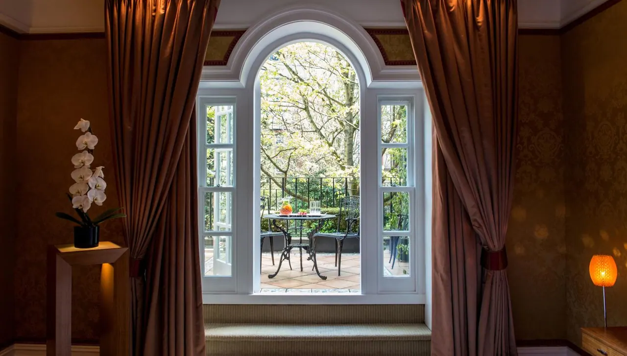 Dark curtains frame an arched window that looks out onto a tiled terrace with a metal bistro table, chairs, railings and trees beyond.