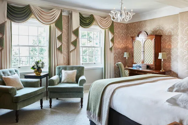 A bedroom with a large bed, two green armchairs, sash windows with decorative drapes, a wooden cabinet and a crystal chandelier.