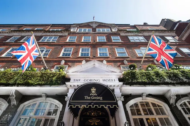 A low-angle view of the brick facade of The Goring Hotel, with its entrance canopy, two Union Jack flags and hedges along the ground floor under a clear blue sky.