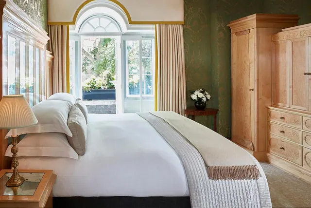 A bedroom with a made double bed, wooden wardrobe and dresser, and French doors opening onto a garden with trees.