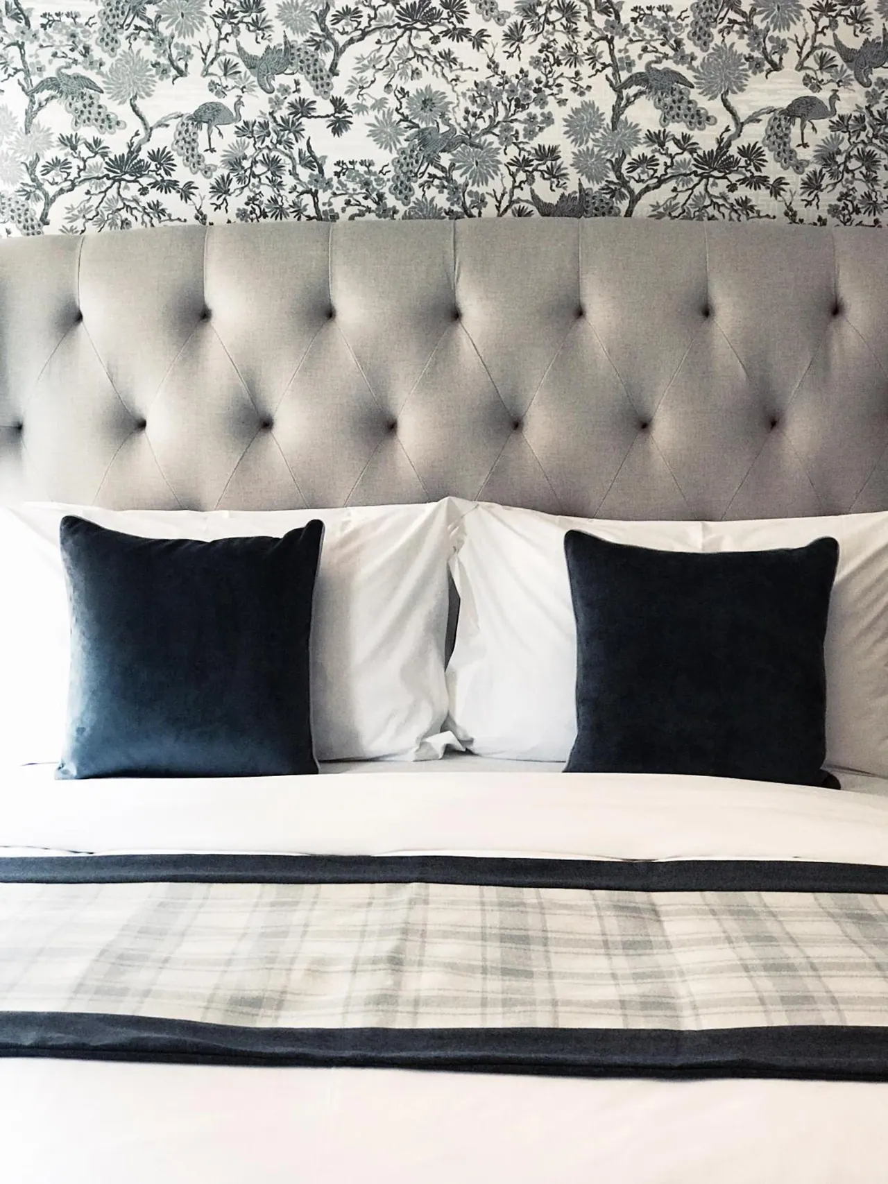 A made bed with a grey tufted headboard, navy velvet cushions and a plaid throw, in front of patterned bird and floral wallpaper.