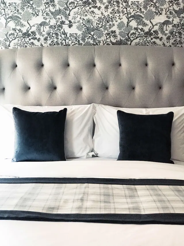A made bed with a grey tufted headboard, navy velvet cushions, white linens and a plaid throw, in front of bird-patterned wallpaper.