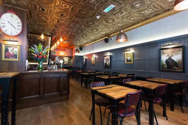 A bar and dining area with a patterned tin ceiling, wooden tables with red leather chairs, a wall clock and framed dog portraits on the walls.