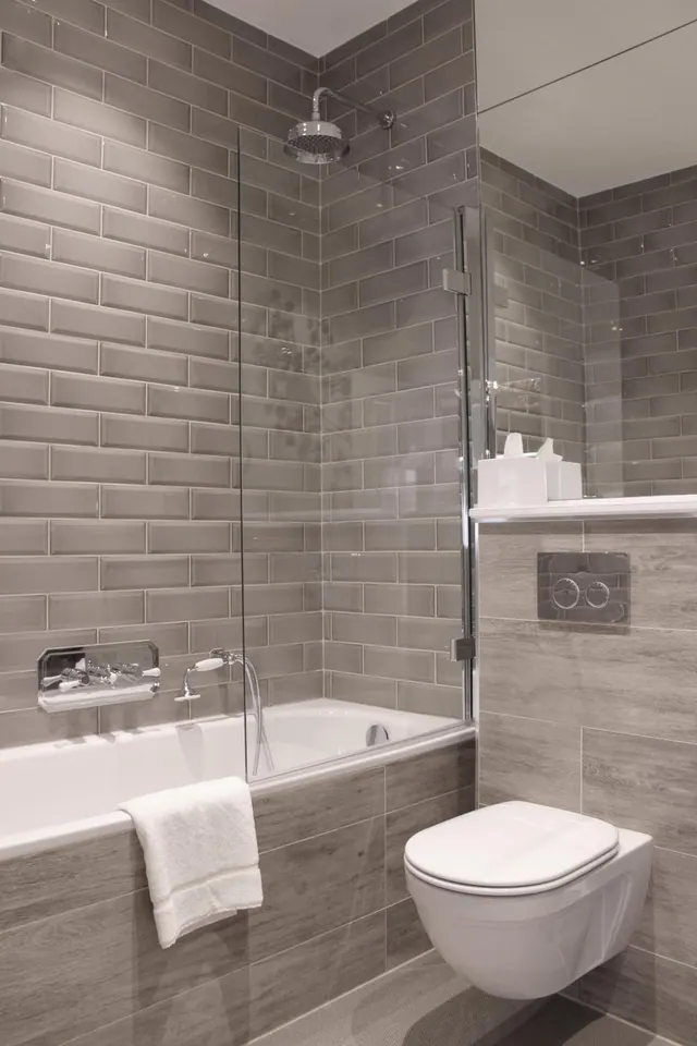 A grey tiled bathroom with a bath, glass shower screen, wall-hung toilet and a white towel draped over the bath edge.