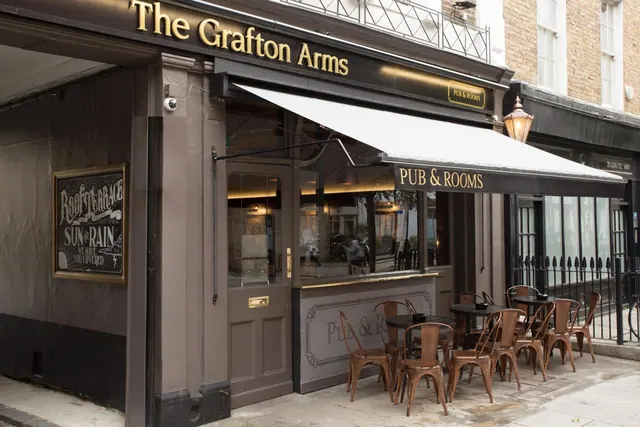The street frontage of The Grafton Arms pub, with a dark awning, gold lettering and metal chairs and tables set out on the pavement.