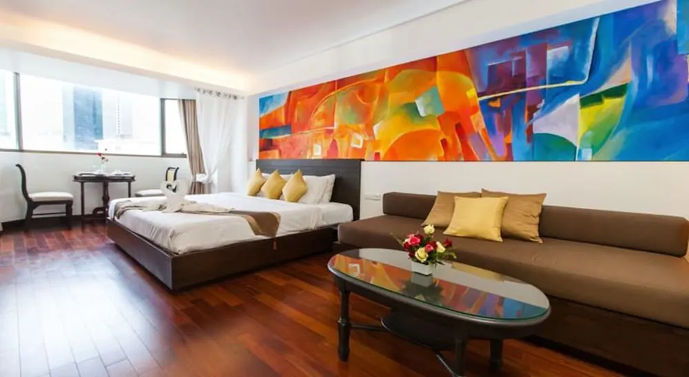 A hotel room with a king bed, a long brown sofa, a glass-topped coffee table and a large colourful abstract painting on the wall.
