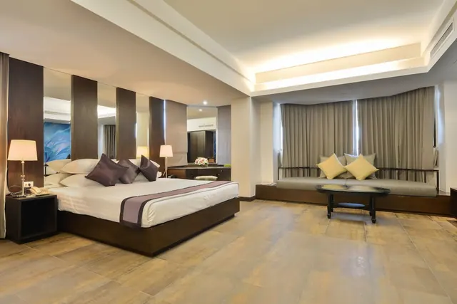 A bedroom with a king bed with purple cushions, a cushioned seating area beside the curtained window, and a low round coffee table on a tiled floor.