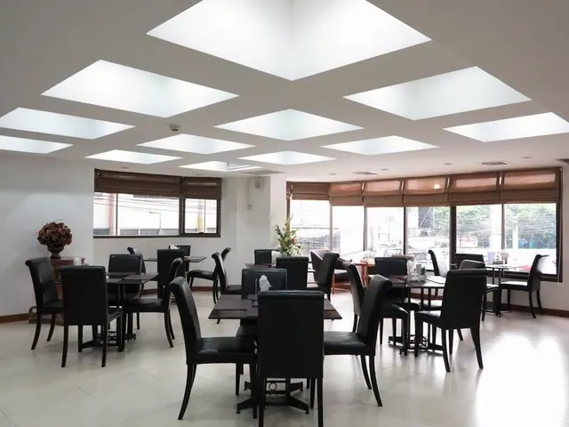 A dining room with black leather chairs and dark tables under a ceiling of skylights, with large windows along the right wall.