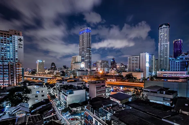 Night view over a dense Bangkok cityscape with illuminated high-rise towers, a road with light trails, and low rooftops in the foreground.