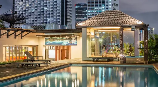 A rooftop swimming pool at night with wooden sun loungers, a glass-walled gym pavilion with a tiled roof, and high-rise buildings behind.
