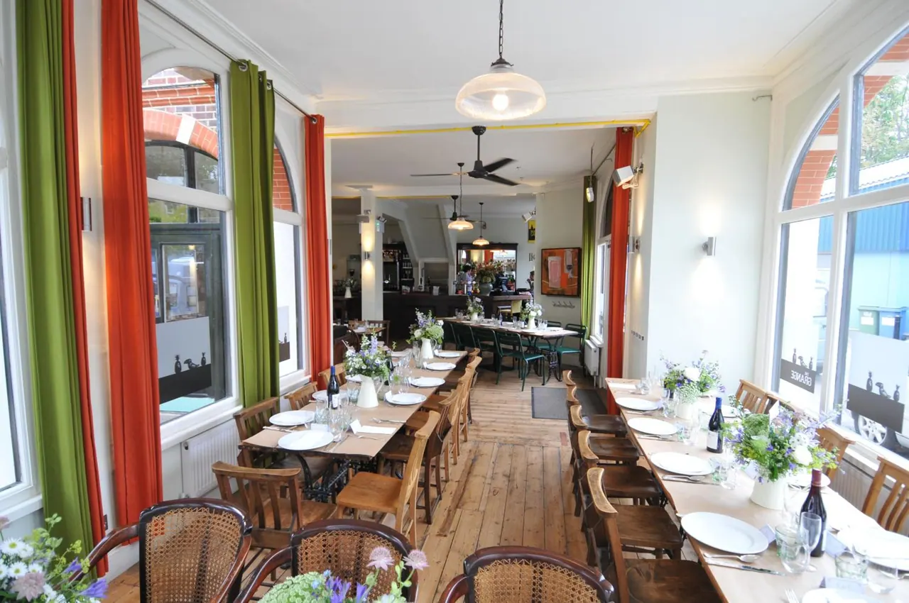A long dining room with set tables, wooden and cane-back chairs, ceiling fans, and tall arched windows framed by green and red curtains.