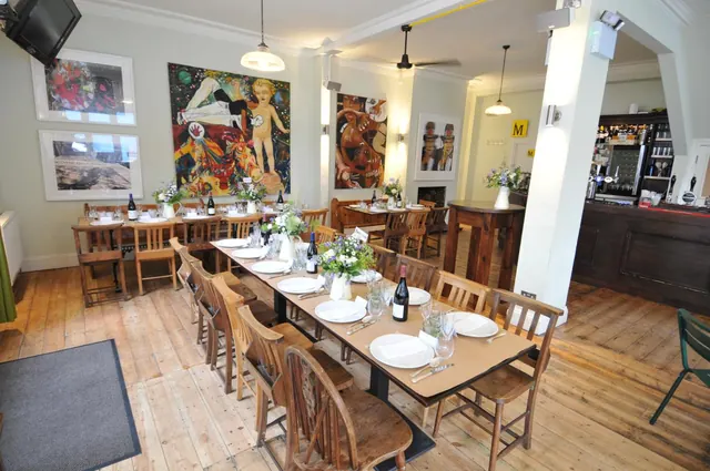 A long wooden dining table is set with white plates, glasses and wine bottles in a restaurant with wooden chairs, large paintings on the walls and a bar to the right.