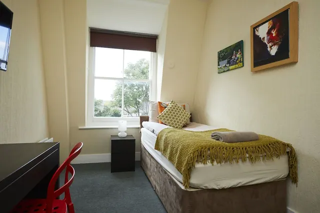 A single bed with an olive throw and cushions sits in a small bedroom beside a window overlooking trees, with a dark desk and red chair on the left.