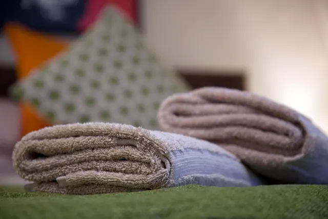 Two rolled beige towels with pale blue trim sit on a green surface in front of blurred green and orange cushions.