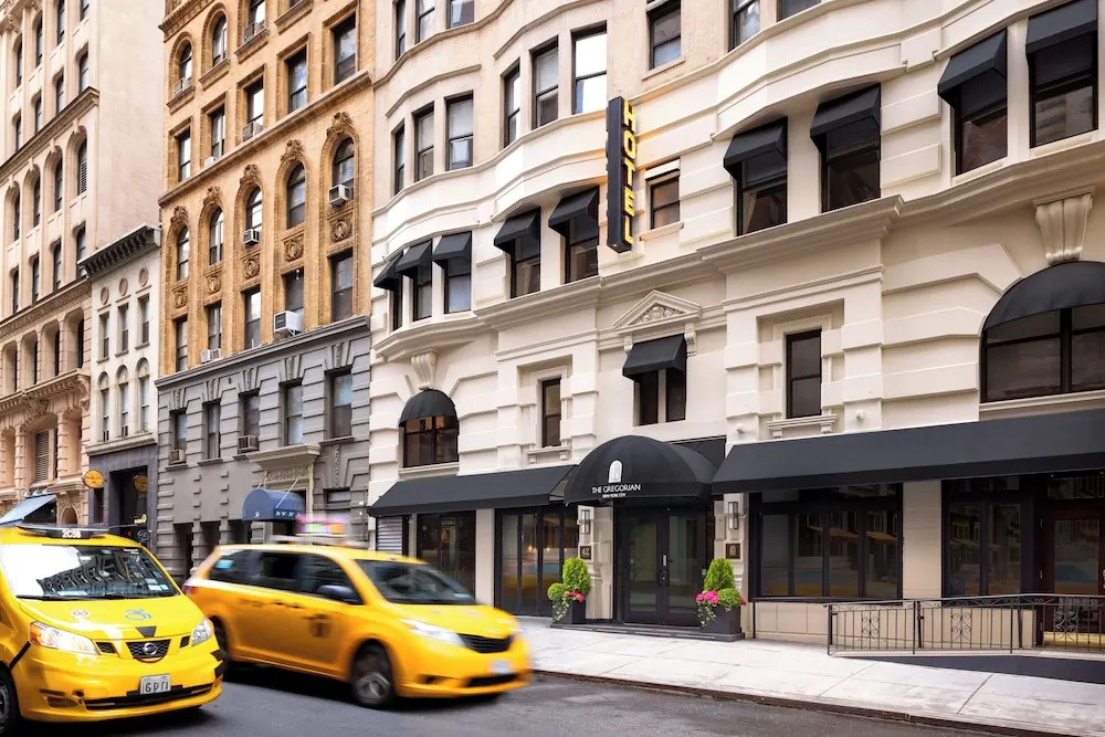 Street-level view of a cream-coloured hotel building with black awnings, with a yellow taxi passing in the foreground.