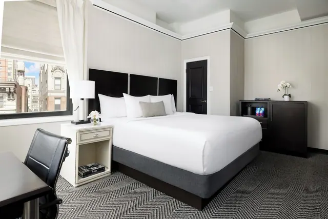 A king bed with white bedding and a black headboard sits in a bedroom beside a window with a view of city buildings.