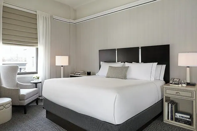 A bedroom with a made king bed with white linens and a dark headboard, two table lamps, and an armchair beside a window with a roman shade.