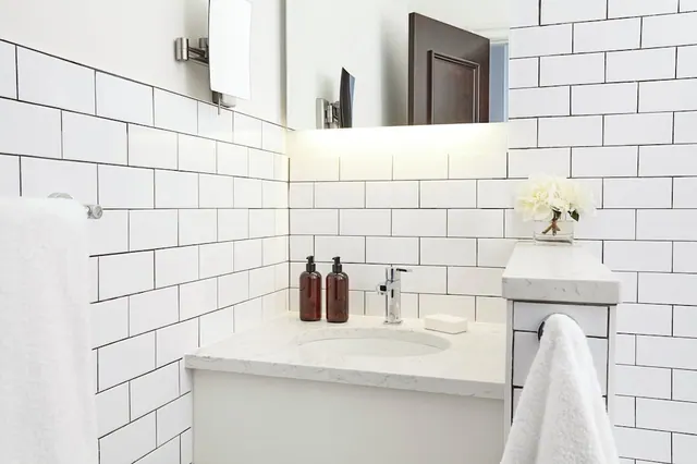 A white subway-tiled bathroom with a quartz-topped vanity, two amber soap dispensers, a flower vase and white hand towels.