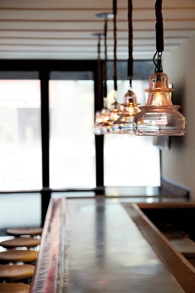 Glass pendant lights hang above a bar counter, with wooden stools along the left and tall windows behind.
