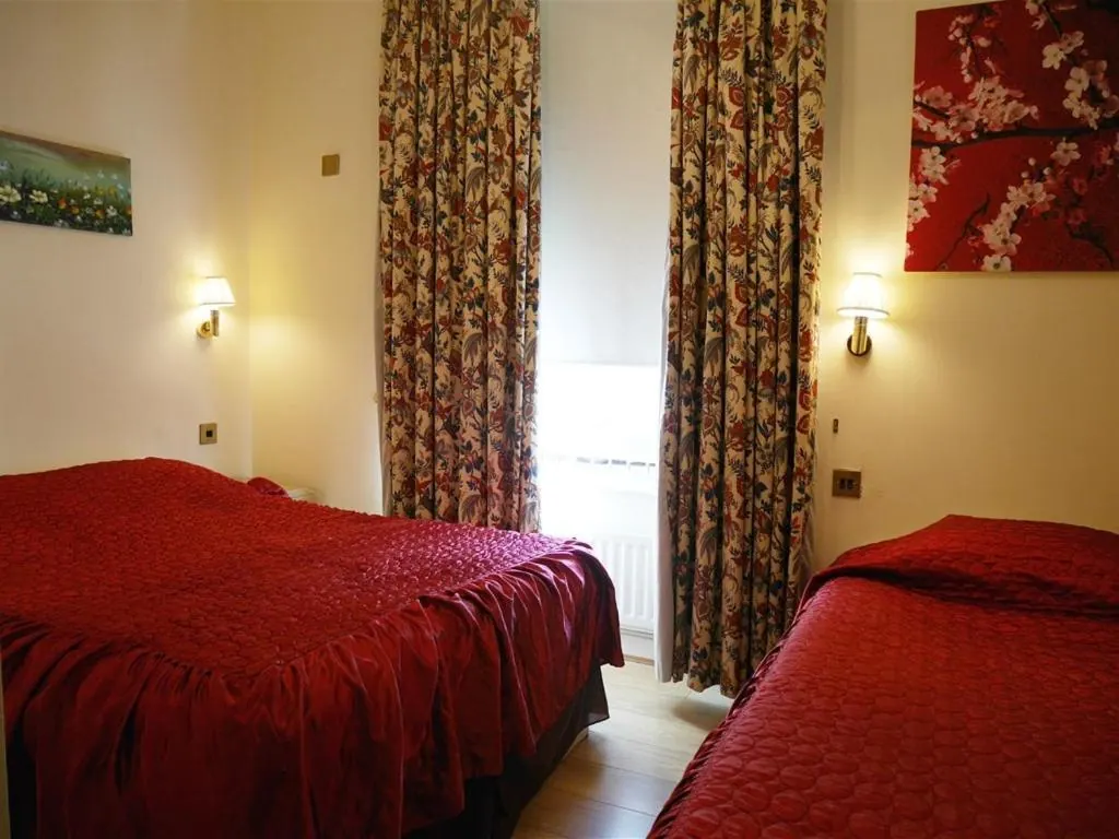 A hotel bedroom with two red quilted beds, floral curtains beside a window, wall lights and a framed picture of red blossoms.