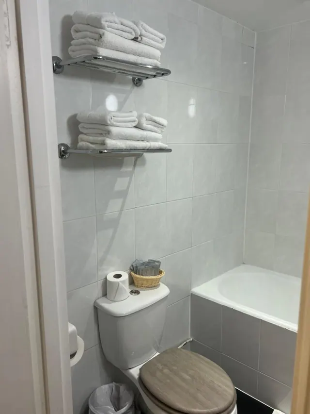 A tiled bathroom with folded white towels on two chrome shelves, a toilet with a wooden seat, a basket of sachets on the cistern, and a bathtub.