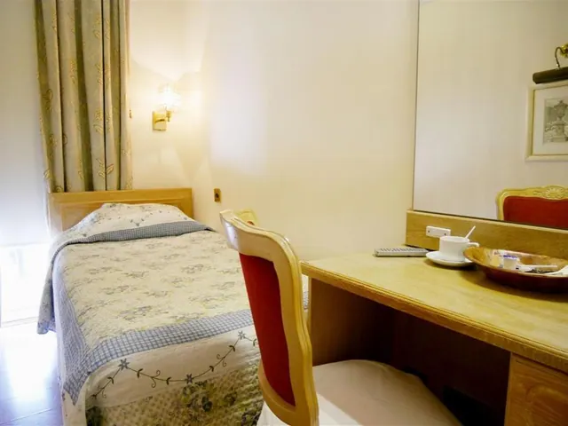 A small hotel bedroom with a single bed, floral curtains, a wall lamp, and a wooden desk with a chair, a cup and saucer, and a mirror.