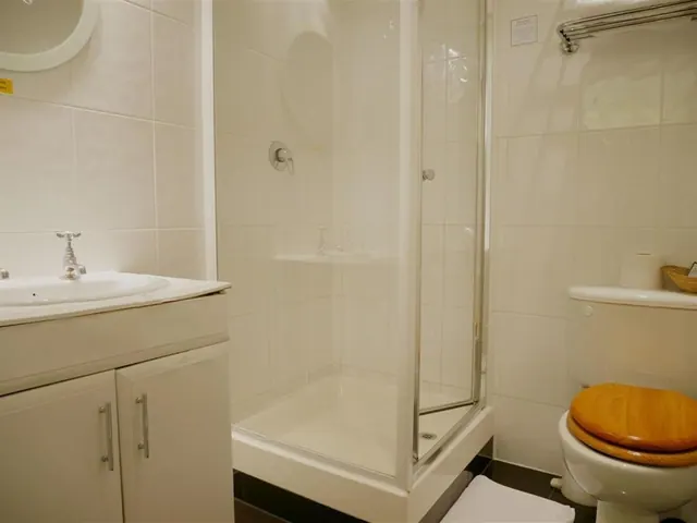 A white tiled bathroom with a pedestal sink, a glass-enclosed shower cubicle and a toilet with a wooden seat.
