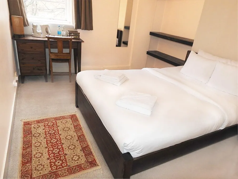 A double bed with white linen and two folded towels sits beside a wooden desk, chair and kettle in a small bedroom.