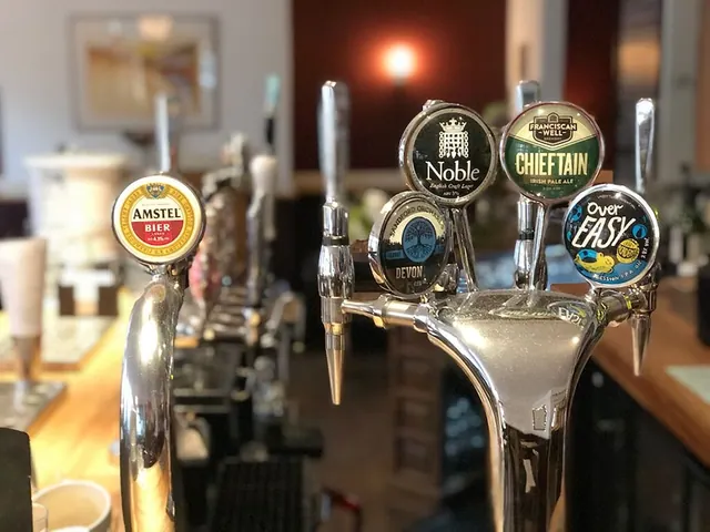 Close-up of several draught beer taps at a bar, with branded badges for Amstel, Noble, Chieftain, Devon and Over Easy, and a blurred room behind.