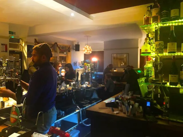A bartender in a dark jumper stands behind a pub bar with beer taps and illuminated bottle shelves, while customers sit in the background.