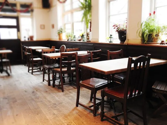 An empty dining room with wooden tables and chairs on a wooden floor, with tall windows and potted plants on the sill.